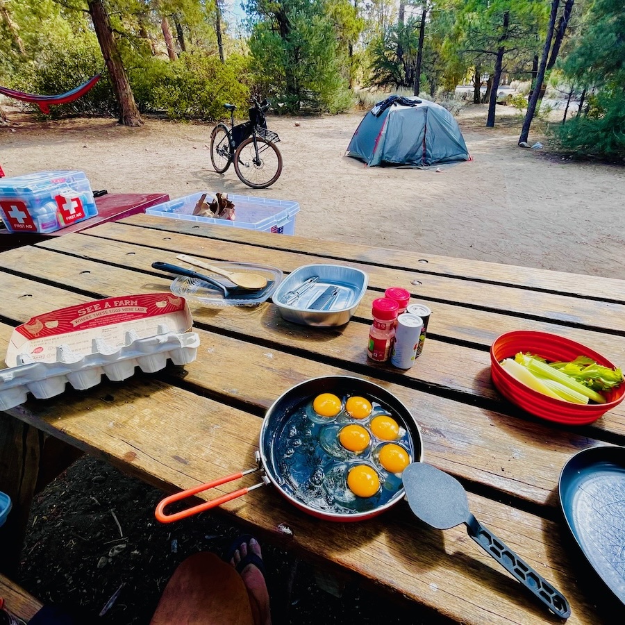 Wooden table with food and cooking supplies at a campsite, with various other outdoor gear and trees in the background.