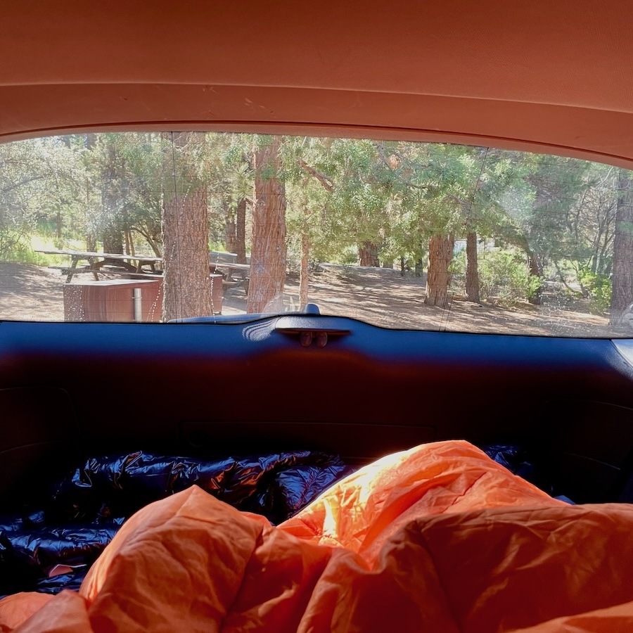 View of a campsite from the rear windshield of a vehicle, with sleeping bags inside the car in the foreground.