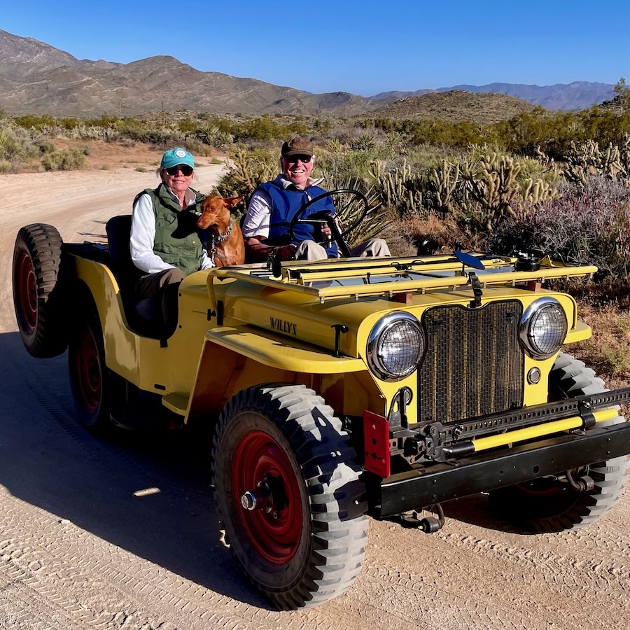 A couple and their dog in front of an older restored open top Jeep in the desert 