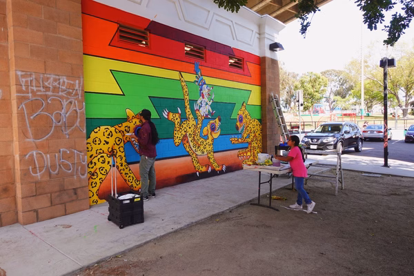 Artist painting a mural on the left while a girl watches and mixes paint on a table to the right