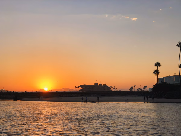 Sunset rising over buildings in the distance, with a beach and water in the foreground
